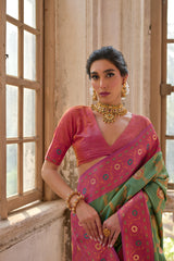 Woman wearing a green silk saree with intricate pink floral zari border and circular motifs, paired with a matching pink blouse and traditional gold jewelry, standing gracefully in a sunlit vintage room with large antique wooden windows and ornate architectural details.