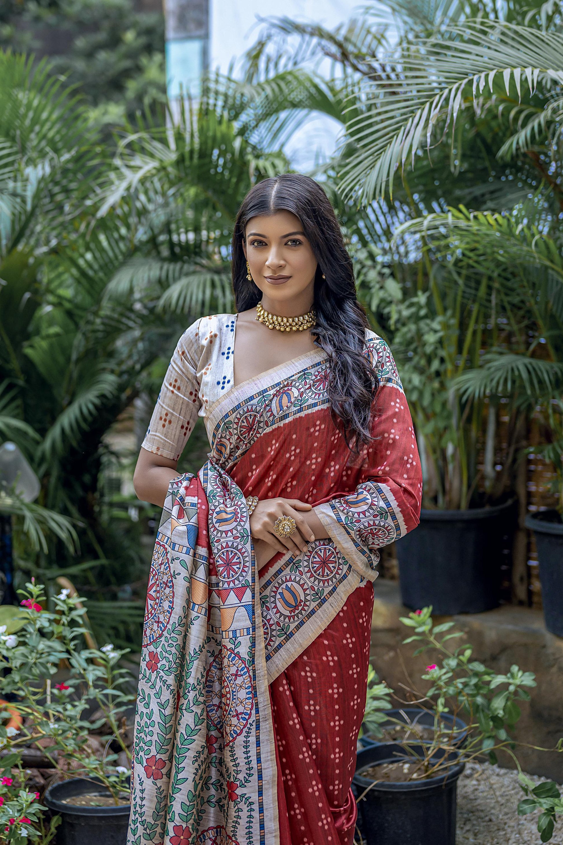 Woman wearing a green saree with intricate white tribal-inspired prints and a multicolor pallu featuring circular and geometric patterns, standing gracefully in a lush outdoor garden surrounded by tropical plants.