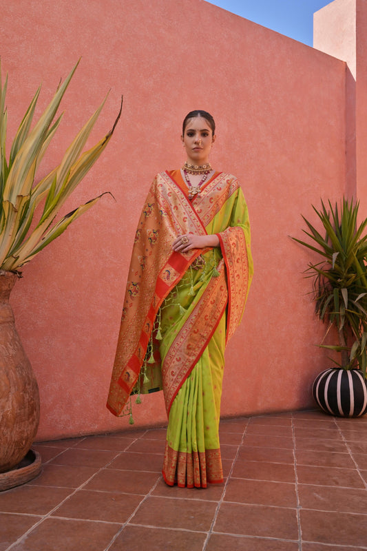 A woman models a vibrant Banarasi Silk Saree. The saree is lime green with fine Zari weaving, featuring a wide, ornate orange and red Zari woven border and pallu. She pairs it with a short-sleeve orange blouse and heavy traditional gold jewelry.