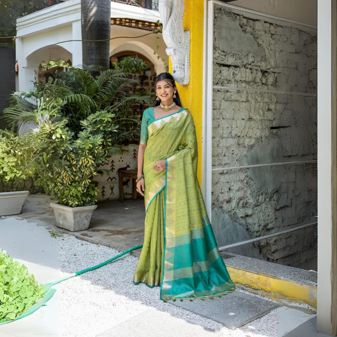 Woman wearing a light green silk saree with gold and teal borders, paired with a matching green blouse and traditional jewelry, standing outdoors near a yellow wall and lush garden plants.