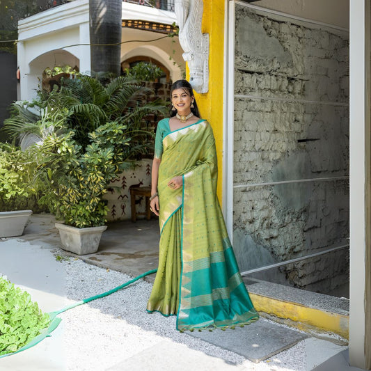 A woman models a vibrant Pista Green Tussar Silk Saree with small woven motifs. The saree has a contrasting peacock blue/teal pallu featuring wide gold Zari stripes. She wears a teal blouse and gold Kundan jewelry, posing in an outdoor courtyard.