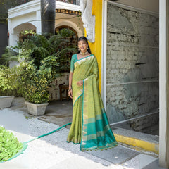 Woman wearing a light green silk saree with gold and teal borders, paired with a matching green blouse and traditional jewelry, standing outdoors near a yellow wall and lush garden plants.