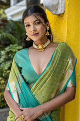 Woman wearing a light green silk saree with gold and teal borders, paired with a matching green blouse and traditional jewelry, standing outdoors near a yellow wall and lush garden plants.