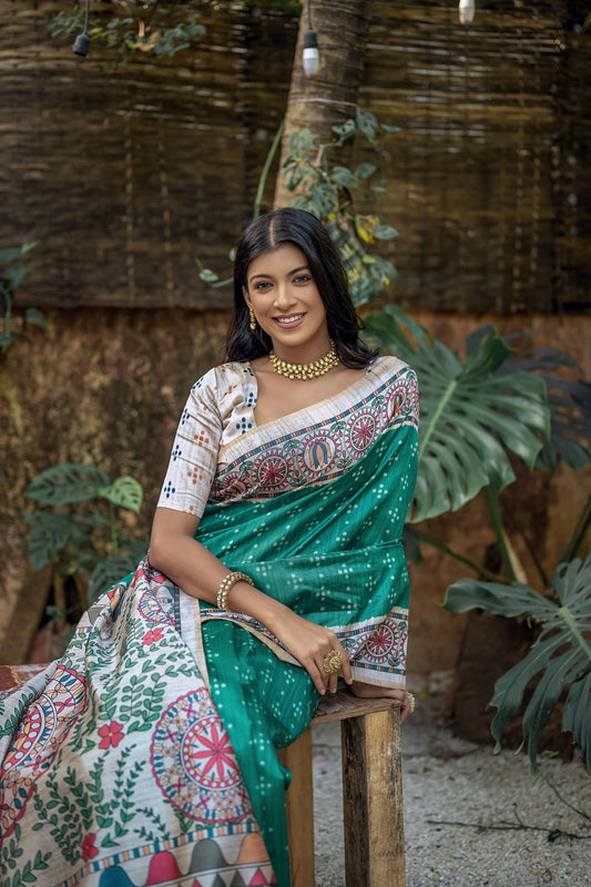 Woman wearing a green saree with intricate white tribal-inspired prints and a multicolor pallu featuring circular and geometric patterns, standing gracefully in a lush outdoor garden with tropical plants and natural sunlight.