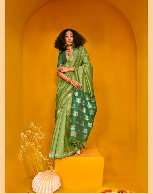 Woman wearing green zari work saree with floral weave and tassel border, paired with ethnic jewellery, standing in a vibrant yellow archway set