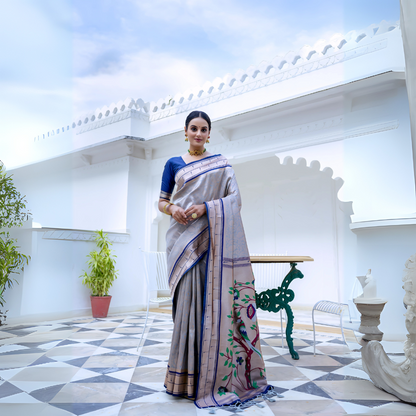 Woman wearing a grey silk saree with blue border and blouse, featuring a handpainted pallu with tree and bird design, posing in a white heritage courtyard with patterned tile flooring and potted plants.