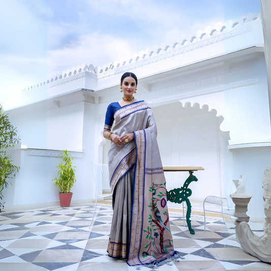 Woman wearing a grey silk saree with blue border and blouse, featuring a handpainted pallu with tree and bird design, posing in a white heritage courtyard with patterned tile flooring and potted plants.