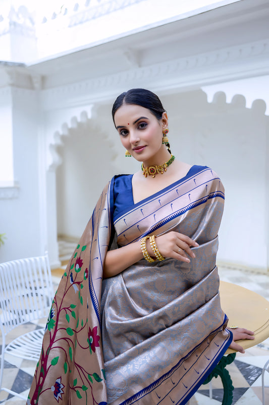 Woman wearing a grey silk saree with blue border and blouse, featuring a handpainted pallu with tree and bird design, posing in a white heritage courtyard with patterned tile flooring and potted plants.
