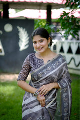 Woman wearing a grey handloom cotton saree with white tribal-inspired prints and a matching patterned blouse, accessorized with silver jewelry, standing on green grass in front of a wall with tribal art motifs in an outdoor garden setting.