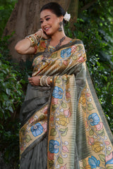 Woman wearing a grey saree with vibrant Madhubani-inspired floral print border and pallu, paired with a matching blouse, standing gracefully on a garden pathway surrounded by lush green trees and foliage.