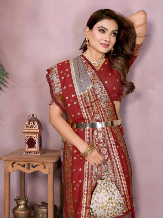 Woman draped in an elegant grey Banarasi saree featuring a rich red and gold embroidered border with intricate traditional motifs, paired with a matching red blouse. She poses gracefully in a modern studio decorated with rustic clay pots, a wooden side table holding brass decor, yellow flowers in a bowl, and a small green branch adding a natural accent to the setting.