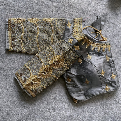 Traditional grey silk blouse featuring intricate gold embroidery and beadwork with floral and paisley motifs on the sleeves and neckline, displayed flat on a textured surface with a bunch of artificial flowers and a woven mat in the background.