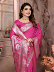 Woman wearing a hot pink silk saree with an ornate silver zari border and intricate pallu motifs, paired with a matching blouse and traditional jewelry, elegantly posing beside a window in a classic floral-themed studio with decorative vases and pastel-toned decor.