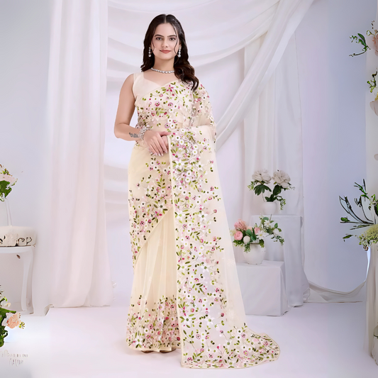 Woman wearing an ivory net saree with intricate pink and green floral embroidery and a matching sleeveless blouse, posing in a modern white studio decorated with draped curtains and fresh floral arrangements