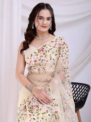 Woman wearing an ivory net saree with intricate pink and green floral embroidery and a matching sleeveless blouse, posing in a modern white studio decorated with draped curtains and fresh floral arrangements