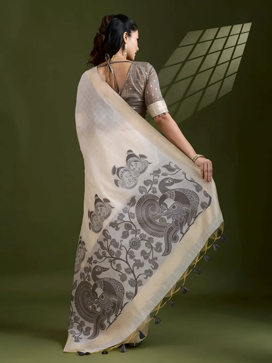 Woman wearing an ivory silk saree with grey fish and floral motifs, paired with a grey blouse and pearl jewelry, posing in a contemporary studio with olive green walls and a metal chair.