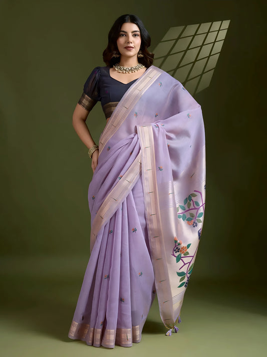 Woman wearing a lavender silk saree with subtle embroidered motifs and a cream-colored border, featuring multicolor floral designs on the pallu, paired with a contrasting black blouse and traditional jewelry, elegantly posing in a studio with olive green walls and a window-patterned light effect.