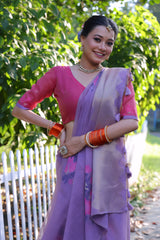 Woman wearing a lavender silk saree with subtle floral embroidery and a contrasting pink blouse, accessorized with traditional jewelry and orange bangles, standing beside a white picket fence in a sunlit garden with lush greenery and trees in the background.
