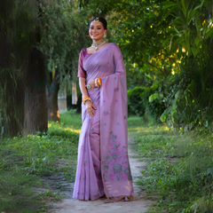  Woman wearing a lavender silk saree with delicate floral embroidery and a matching blouse, accessorized with traditional jewelry and bangles, standing and smiling on a sunlit garden path surrounded by lush green trees and foliage.