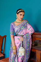 Woman wearing a lavender and pink silk saree with intricate floral and geometric borders, paired with a matching blouse and traditional jewelry, elegantly posing in a vintage-inspired room with teal blue walls, antique wooden furniture, and decorative ceramic accents.