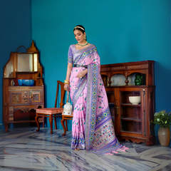 Woman wearing a lavender and pink silk saree with intricate floral and geometric borders, paired with a matching blouse and traditional jewelry, elegantly posing in a vintage-inspired room with teal blue walls, antique wooden furniture, and decorative ceramic accents.