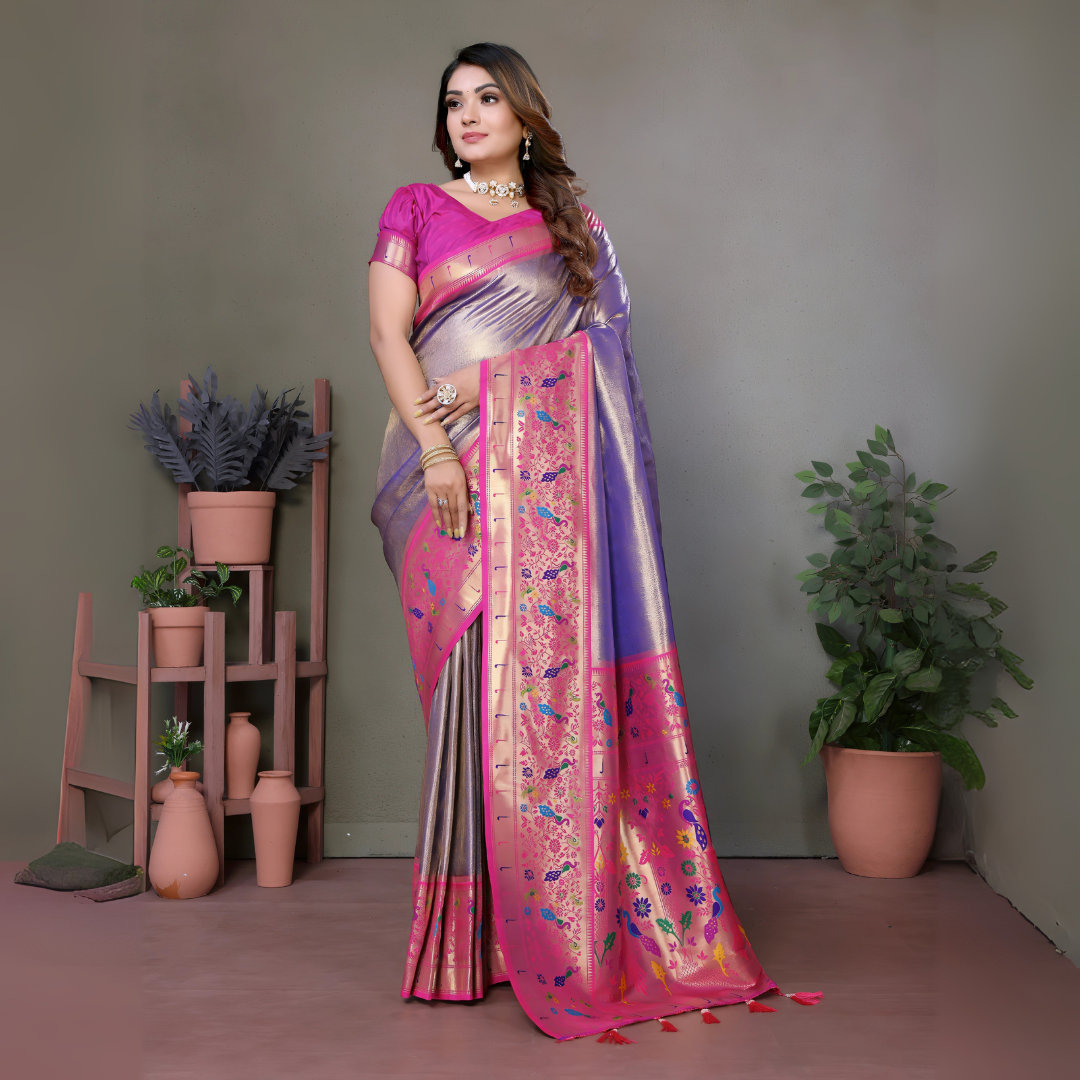 Woman wearing a lavender and pink silk saree with a rich golden border and multicolor floral motifs, paired with a magenta blouse and traditional jewelry. She poses gracefully in a modern studio setting with minimalist decor, including potted plants and terracotta vases on wooden stands against a muted background.