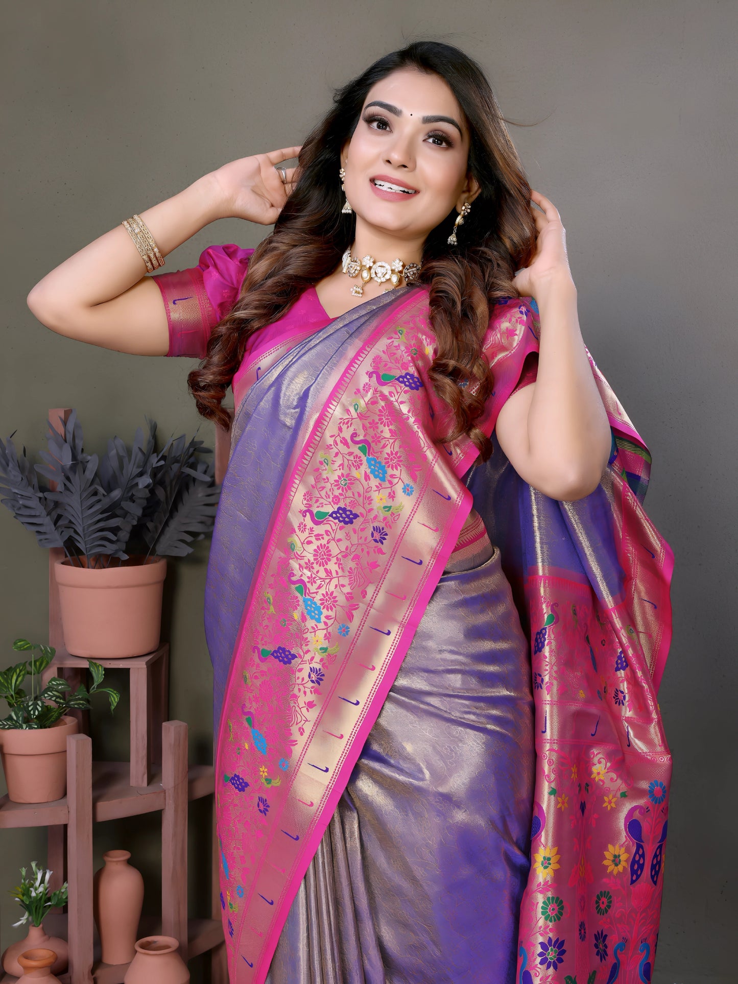 Woman wearing a lavender and pink silk saree with a rich golden border and multicolor floral motifs, paired with a magenta blouse and traditional jewelry. She poses gracefully in a modern studio setting with minimalist decor, including potted plants and terracotta vases on wooden stands against a muted background.