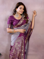 Woman wearing a lavender and purple Banarasi silk saree with an ornate golden border and intricate woven pallu featuring traditional motifs. She pairs the saree with a matching purple blouse adorned with gold detailing and statement jewelry, posing gracefully in a modern studio setting with potted green plants and blush-toned vases in the background.