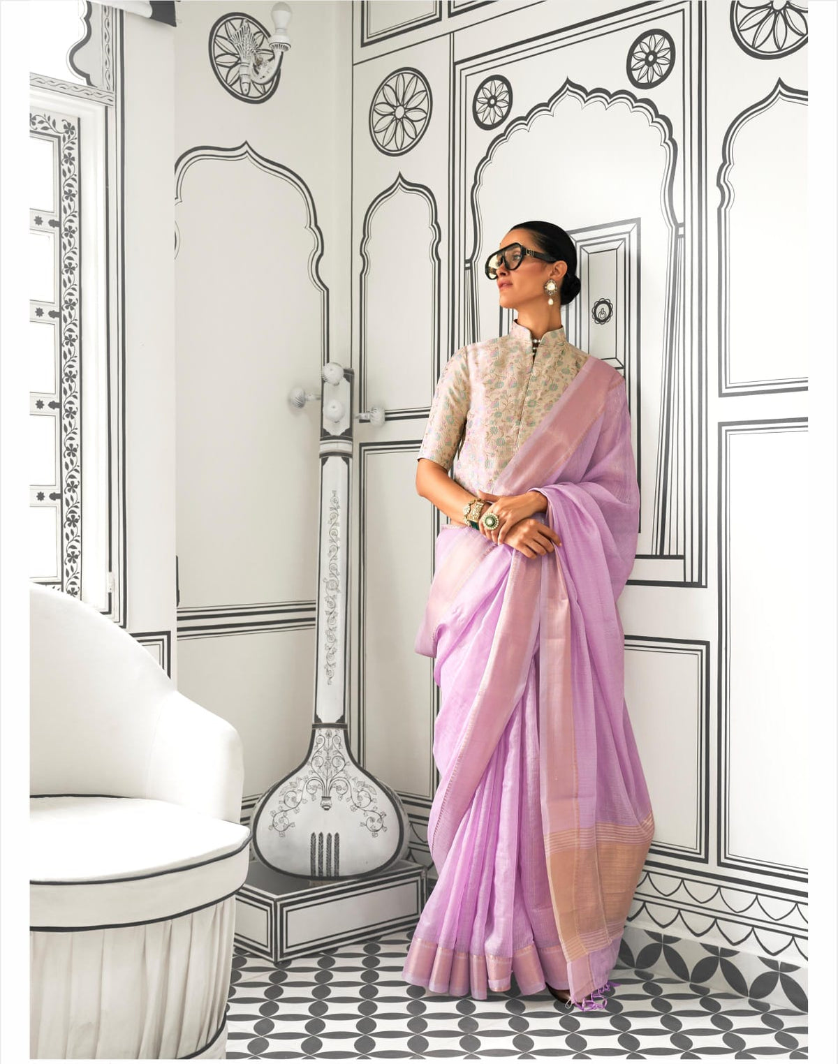 Woman in a lavender saree with zari work standing in a room with intricate black-and-white wall illustrations and a tanpura drawing