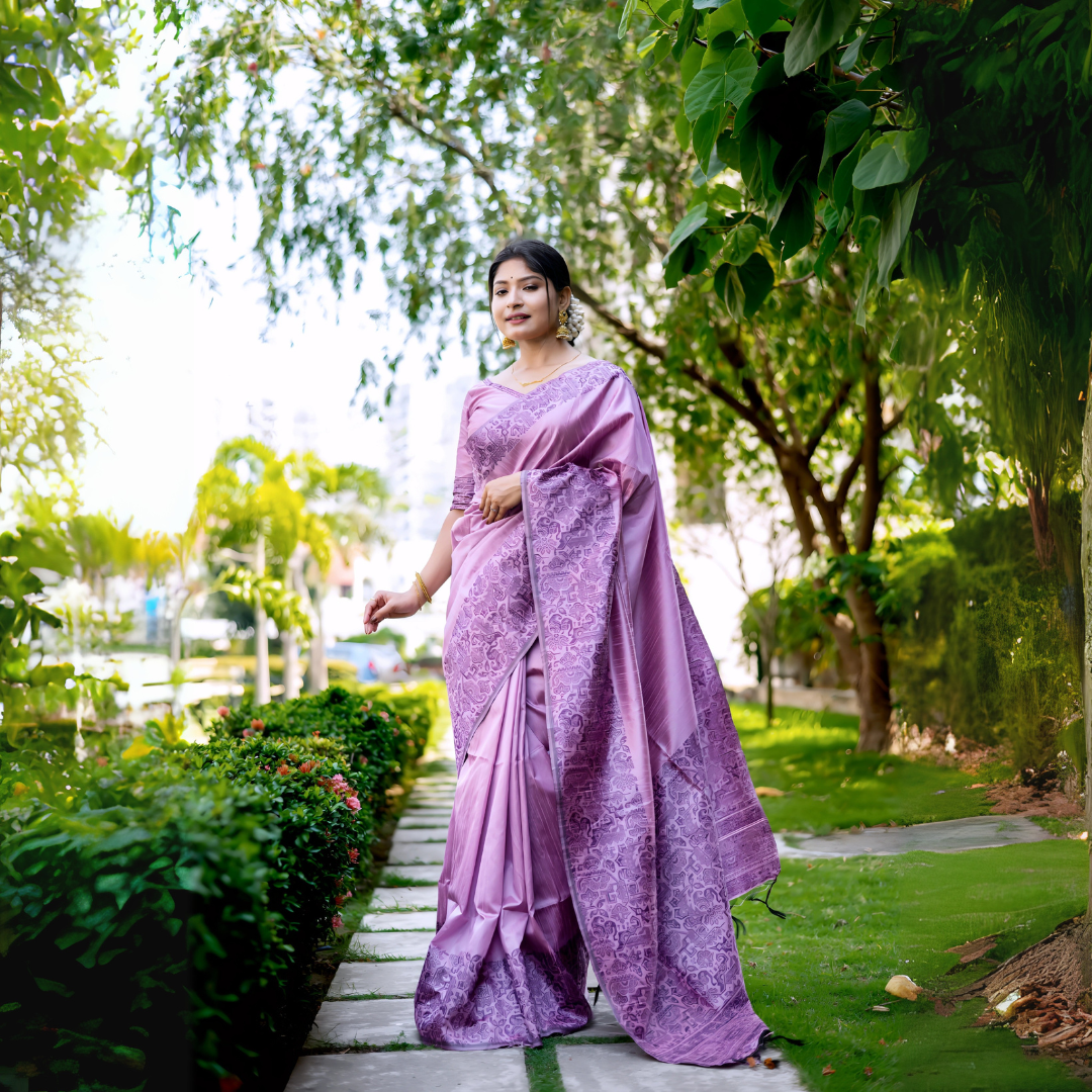 Woman wearing a lavender silk saree with intricate floral patterns, paired with a matching blouse and statement earrings, elegantly walking along a stone pathway in a lush green garden with trees and manicured lawns.