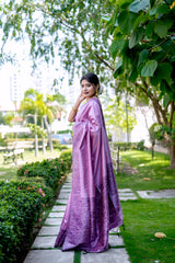 Woman wearing a lavender silk saree with intricate floral patterns, paired with a matching blouse and statement earrings, elegantly walking along a stone pathway in a lush green garden with trees and manicured lawns.