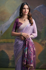 Woman wearing a lavender silk saree with intricate peacock embroidery and floral motifs on the pallu, paired with a matching blouse and traditional jewelry, standing in front of a painted mountain and forest backdrop.