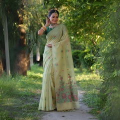  Woman wearing a light yellow silk saree with pastel floral embroidery and a green blouse, accessorized with traditional jewelry and bangles, standing on a sunlit garden path surrounded by lush green trees and foliage