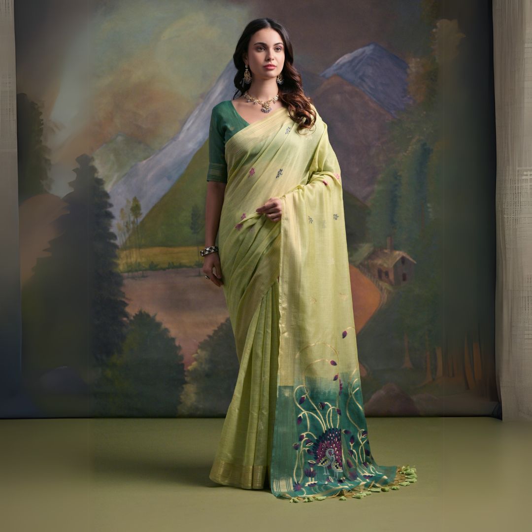 Woman wearing a light green silk saree with delicate floral embroidery and a prominent peacock motif on the pallu, paired with a contrasting green blouse and traditional jewelry, standing in front of a painted mountain and forest backdrop