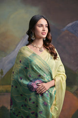 Woman wearing a light green silk saree with delicate floral embroidery and a prominent peacock motif on the pallu, paired with a contrasting green blouse and traditional jewelry, standing in front of a painted mountain and forest backdrop.