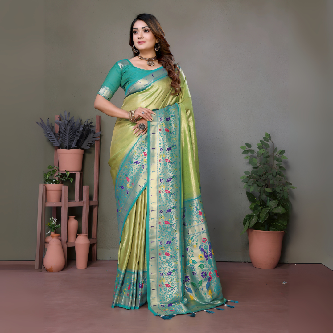 Woman wearing a lime green and blue silk saree with a floral-patterned pallu and pale turquoise border, paired with a contrasting teal blouse. She is accessorized with a choker necklace, bangles, and rings, posing elegantly in a modern studio with potted plants, terracotta vases, and minimalist wooden stands against a muted backdrop.