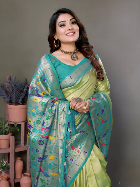Woman wearing a lime green and blue silk saree with a floral-patterned pallu and pale turquoise border, paired with a contrasting teal blouse. She is accessorized with a choker necklace, bangles, and rings, posing elegantly in a modern studio with potted plants, terracotta vases, and minimalist wooden stands against a muted backdrop.