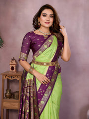 Woman wearing a lime green Banarasi silk saree with a rich purple and gold embroidered border, featuring intricate traditional motifs and an ornate pallu design. She pairs the saree with a matching purple blouse adorned with gold detailing, accessorized with traditional jewelry and bangles, posing gracefully in a modern studio setting with large white vases, a wooden side table, brass decor, green foliage, and a bowl of yellow flowers.