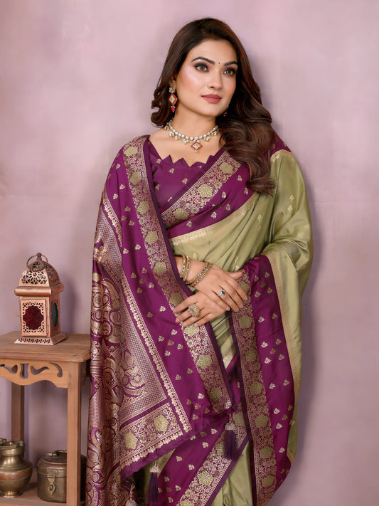 Woman wearing a lime green Banarasi silk saree with a rich purple and gold embroidered border, featuring intricate traditional motifs and an ornate pallu design. She pairs the saree with a matching purple blouse adorned with gold detailing, accessorized with traditional jewelry and bangles, posing gracefully in a modern studio setting with large white vases, a wooden side table, brass decor, green foliage, and a bowl of yellow flowers.