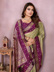 Woman wearing a lime green Banarasi silk saree with a rich purple and gold embroidered border, featuring intricate traditional motifs and an ornate pallu design. She pairs the saree with a matching purple blouse adorned with gold detailing, accessorized with traditional jewelry and bangles, posing gracefully in a modern studio setting with large white vases, a wooden side table, brass decor, green foliage, and a bowl of yellow flowers.