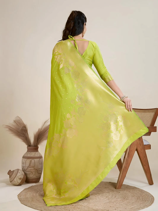 Woman wearing a lime green silk saree with a matching blouse and gold jewelry, elegantly posing beside a wooden chair in a minimalist studio setting with neutral decor, ceramic vases, and pampas grass.