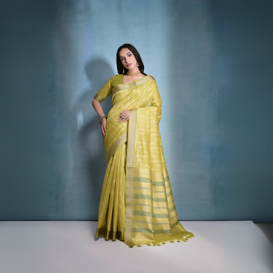 Woman wearing a lime yellow silk saree with subtle stripes and patterned border, paired with a matching blouse and traditional gold jewelry, posing gracefully in a blue-gradient studio backdrop.