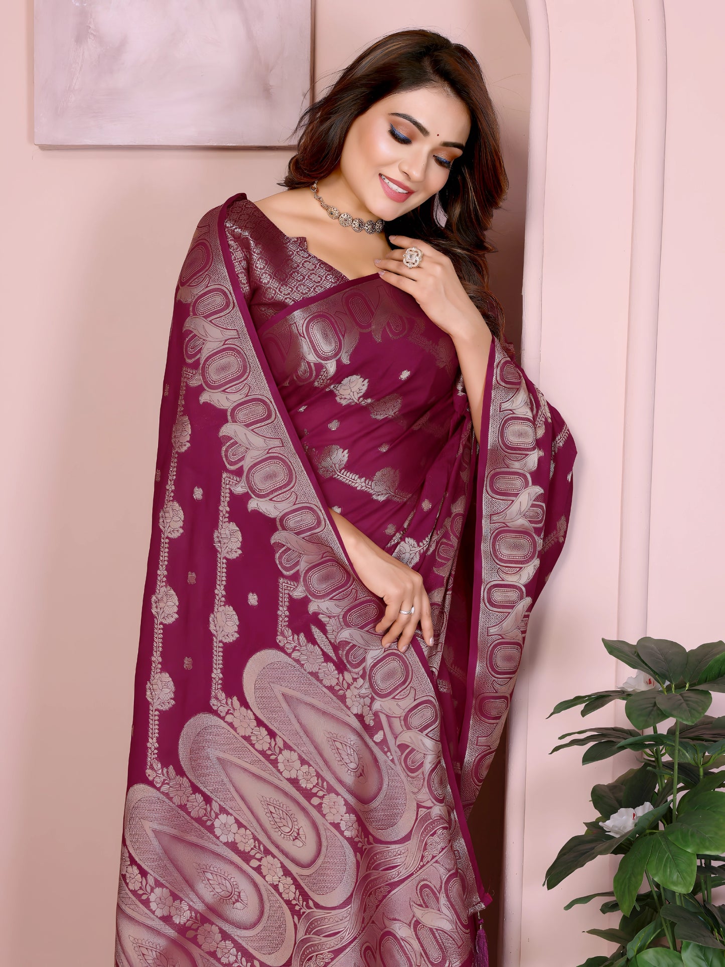 Woman wearing a magenta Banarasi saree with intricate silver zari motifs and rich ethnic border, paired with a matching silver blouse with short sleeves. She poses gracefully in an elegant arched pastel setting with minimal decor, including a white pedestal with vases and a potted green plant, showcasing a traditional Indian festive look.
