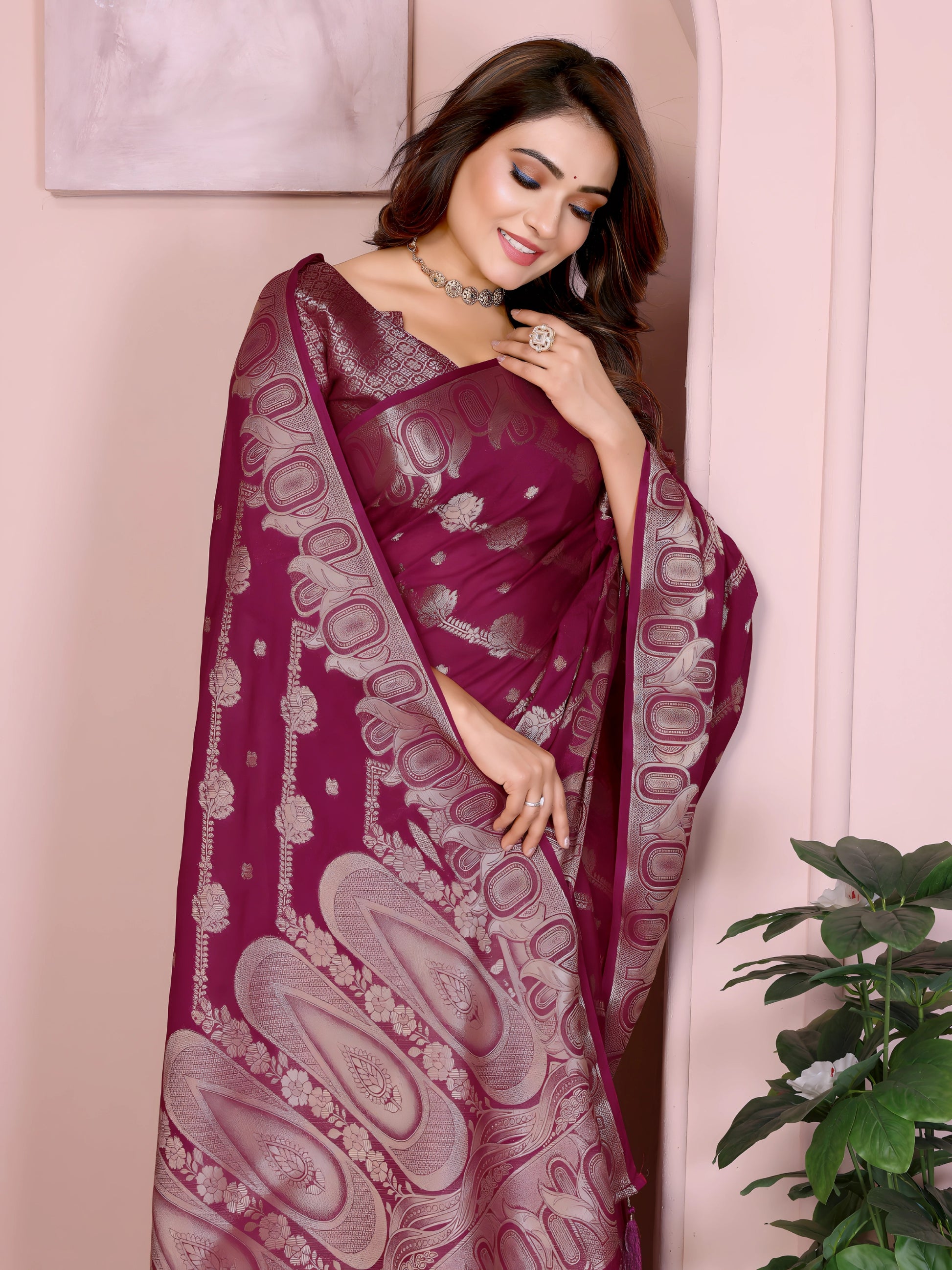 Woman wearing a magenta Banarasi saree with intricate silver zari motifs and rich ethnic border, paired with a matching silver blouse with short sleeves. She poses gracefully in an elegant arched pastel setting with minimal decor, including a white pedestal with vases and a potted green plant, showcasing a traditional Indian festive look.