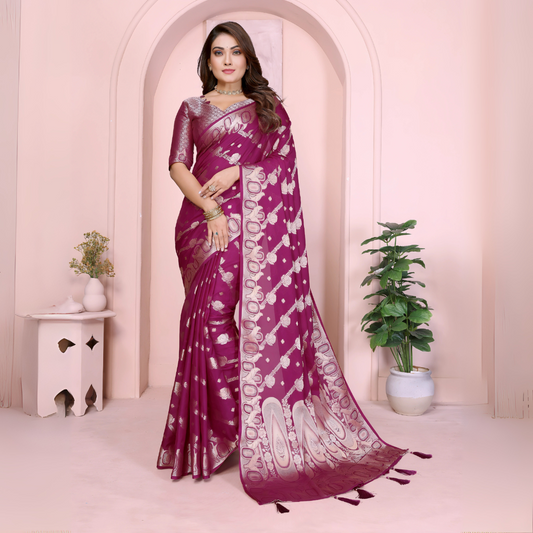Woman wearing a magenta Banarasi saree with intricate silver zari motifs and rich ethnic border, paired with a matching silver blouse with short sleeves. She poses gracefully in an elegant arched pastel setting with minimal decor, including a white pedestal with vases and a potted green plant, showcasing a traditional Indian festive look.