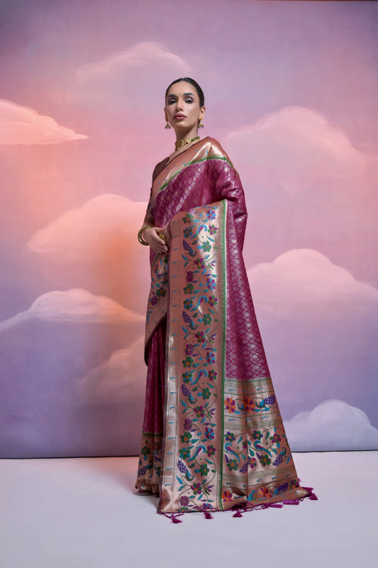 Woman wearing a magenta Banarasi silk saree with intricate gold and multicolor floral zari border and pallu, paired with a matching blouse and traditional jewelry, posing in a pastel-themed studio with soft cloud-like background.