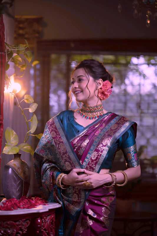 Woman wearing a magenta Banarasi silk saree with intricate gold and teal blue border, paired with a teal blue blouse and traditional jewelry, standing beside an ornate red mirror and console table in a vintage-inspired interior with checkered flooring and wooden blinds.