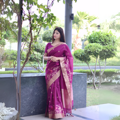 Woman wearing a magenta saree with beige floral embroidery and border, posing outdoors near green landscaped bushes and trees.