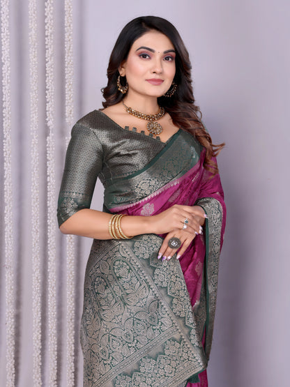 Woman wearing a magenta Banarasi silk saree with a silver and grey zari border, intricate woven motifs, and a matching grey blouse. She is adorned with traditional jewelry and poses gracefully in a modern studio setting featuring woven baskets filled with rose petals and white curtain drapes against a neutral backdrop.