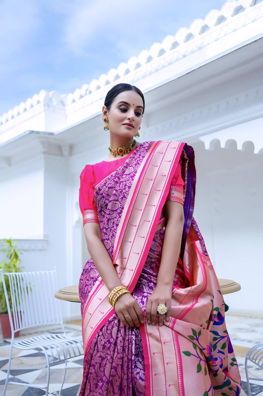 Woman wearing a magenta silk saree with intricate woven patterns and a contrasting peach pallu featuring handpainted floral and bird designs, styled with a matching blouse and jewelry, posing in a white heritage courtyard with arched walls and patterned tile flooring